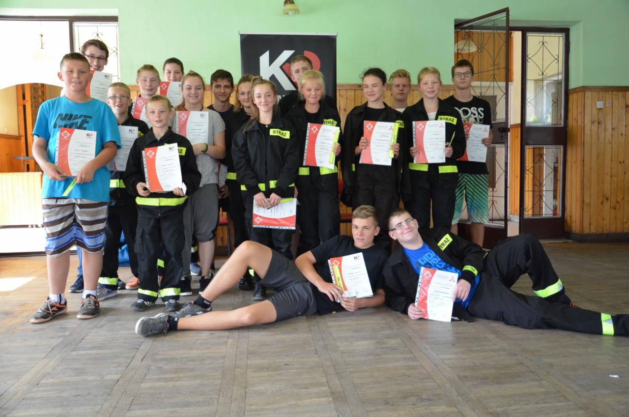 Grupa nastolatków i młodych dorosłych, ubranych w stroje strażackie i codzienne ubrania, pozuje z certyfikatami na tle sali z drewnianą podłogą i zielonymi ścianami, po ukończonym szkoleniu.