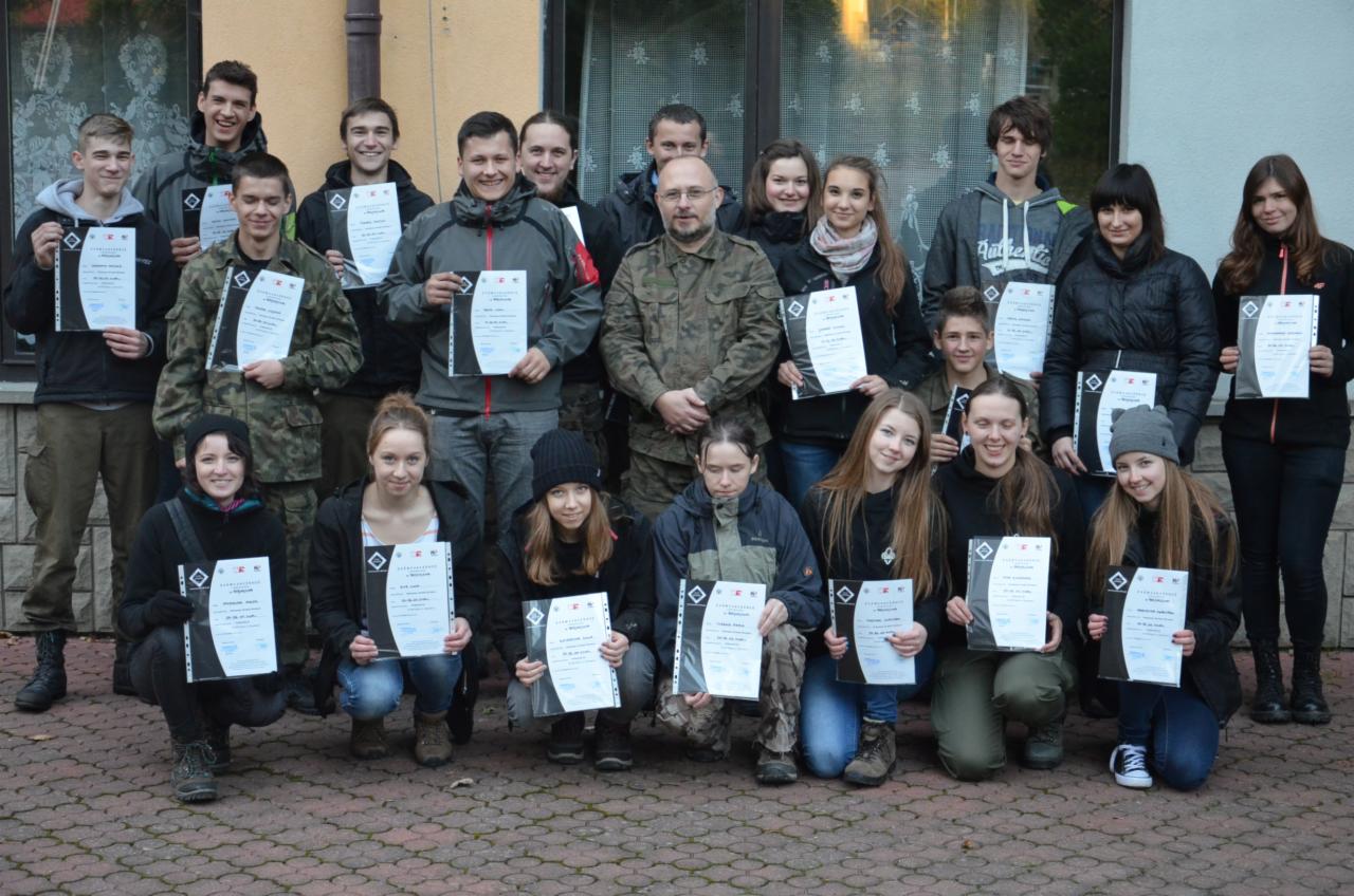 Grupa młodych ludzi, w tym jedna osoba w mundurze wojskowym, pozuje do zdjęcia trzymając certyfikaty na zewnątrz budynku.