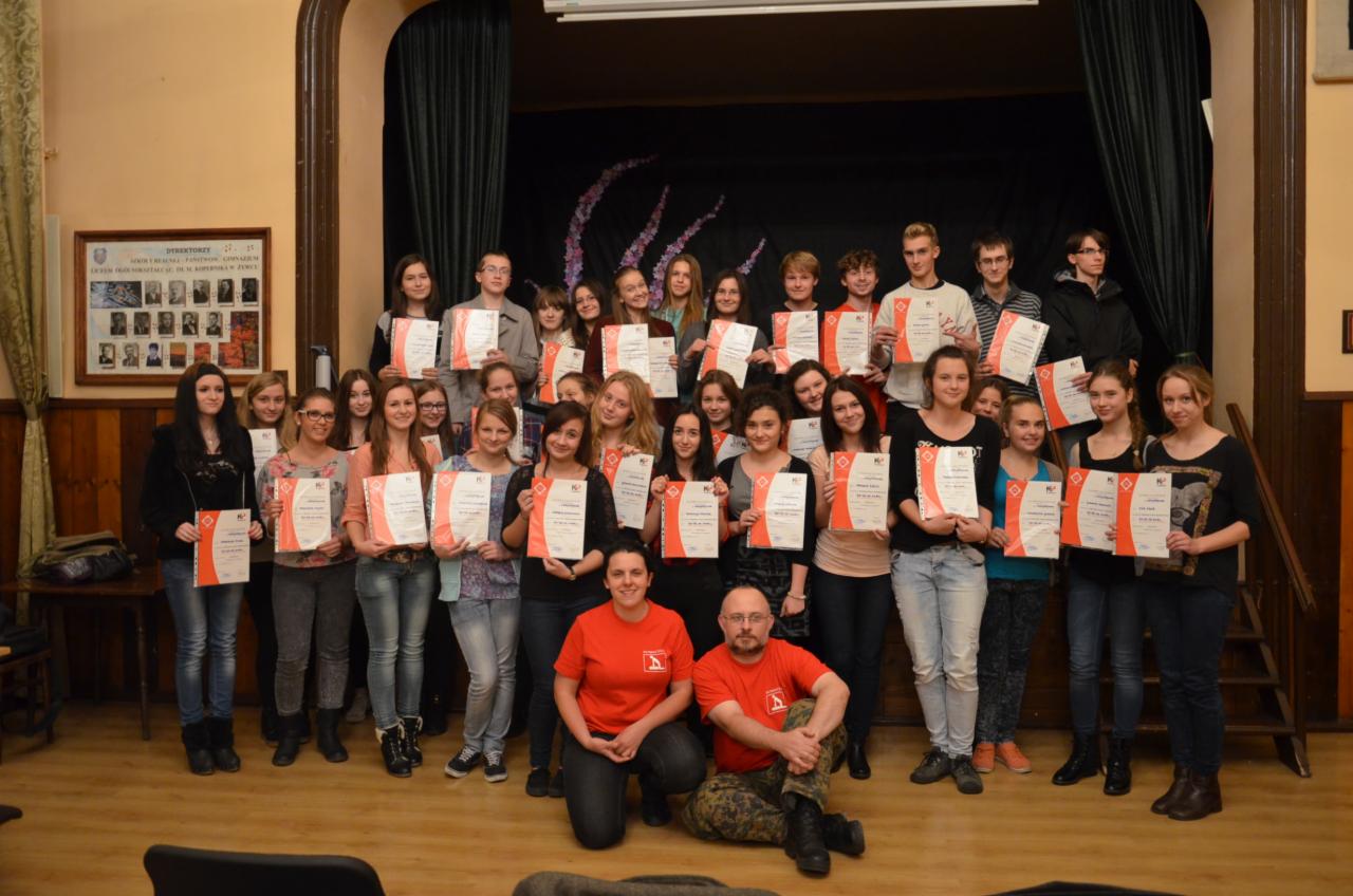 Grupa młodych osób, w tym dwóch instruktorów w czerwonych koszulkach, pozuje na scenie trzymając certyfikaty ukończenia kursu pierwszej pomocy, z czarnym tłem i dekoracjami w kształcie gałęzi.