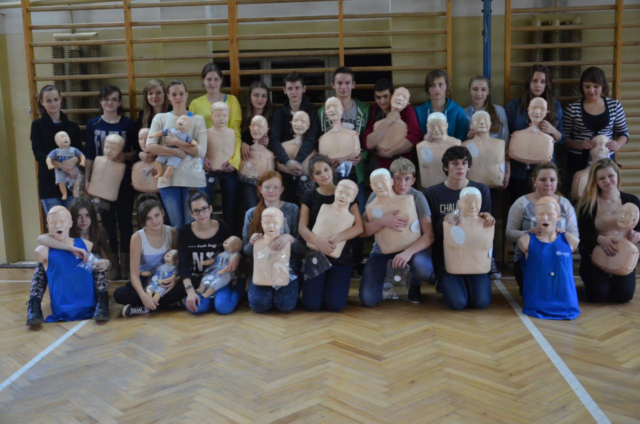 Grupa nastolatków pozuje z manekinami do resuscytacji na tle drewnianej sali gimnastycznej, niektórzy trzymają dodatkowo lalki niemowlęce.
