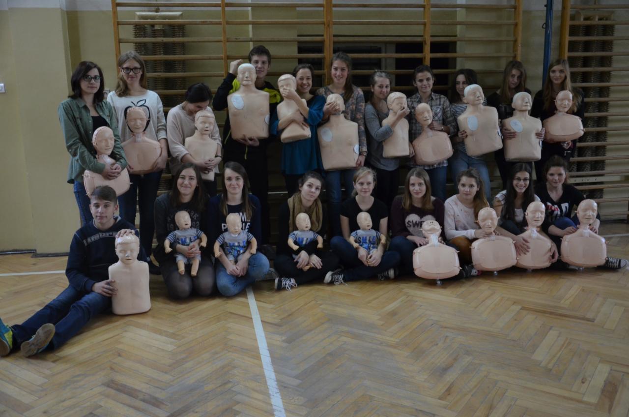 Grupa młodych ludzi, stojących i siedzących na parkiecie w sali gimnastycznej, trzymających manekiny do nauki resuscytacji, niektóre w kształcie torsów dorosłych, inne w kształcie niemowląt.