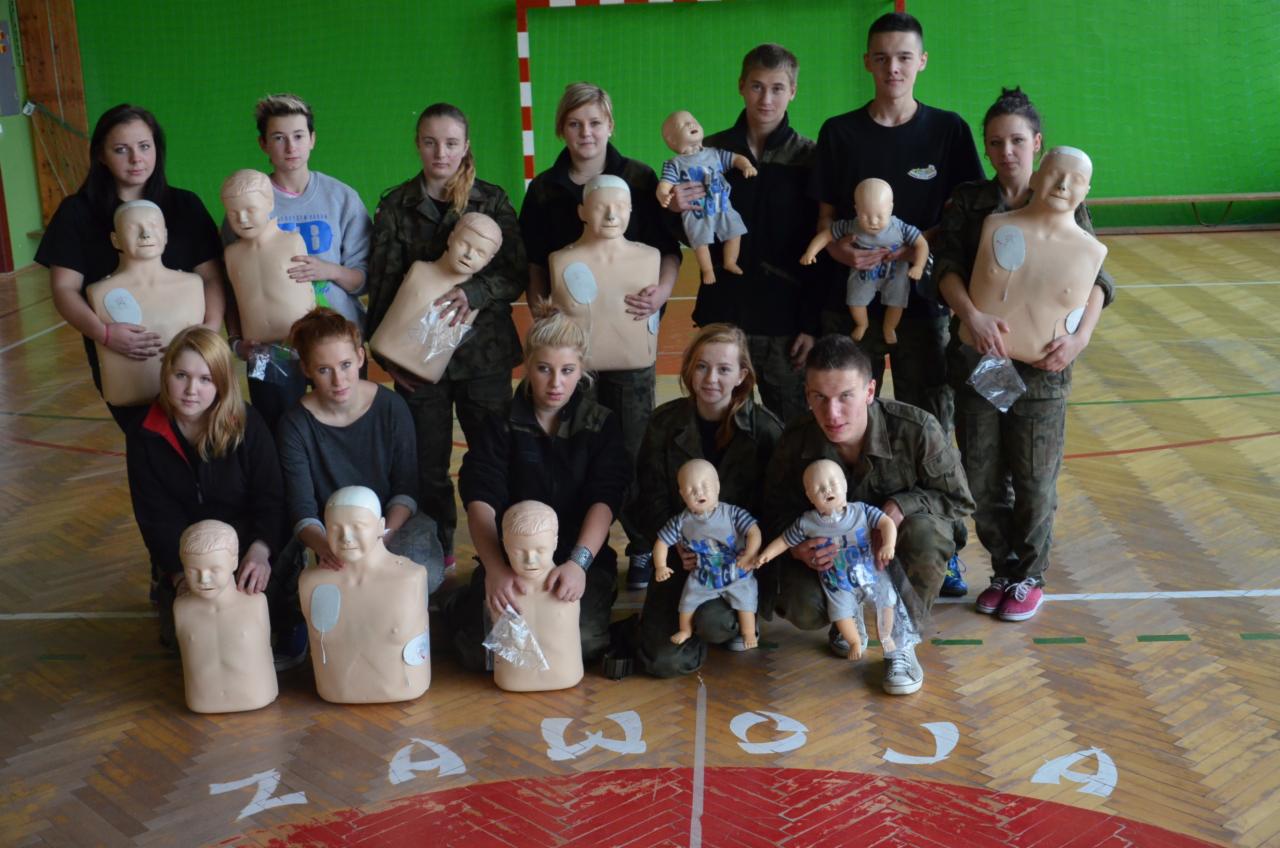Grupa osób, w tym osoby w mundurach, pozuje z fantomami do resuscytacji dorosłych i niemowląt na sali gimnastycznej z napisem na podłodze.