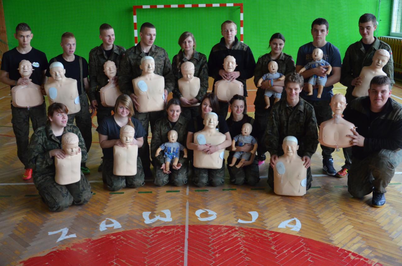 Grupa młodych ludzi, ubranych częściowo w mundury, pozuje w sali gimnastycznej trzymając fantomy do ćwiczeń resuscytacji krążeniowo-oddechowej oraz lalki niemowlęce.