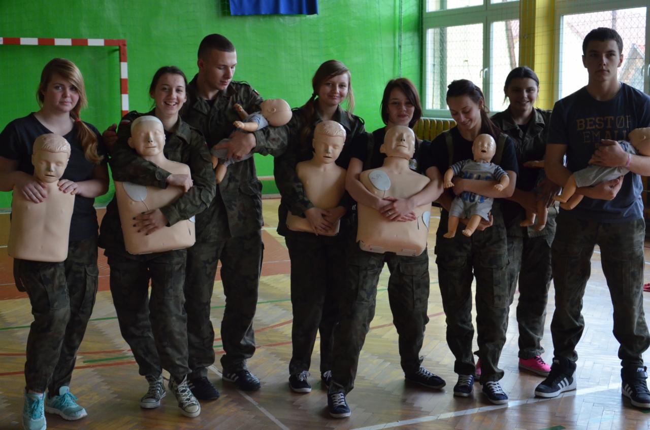 Grupa młodych osób, w tym w mundurach, ćwiczy udzielanie pierwszej pomocy na fantomach i lalkach niemowlęcych w jasnym pomieszczeniu w Żywcu.