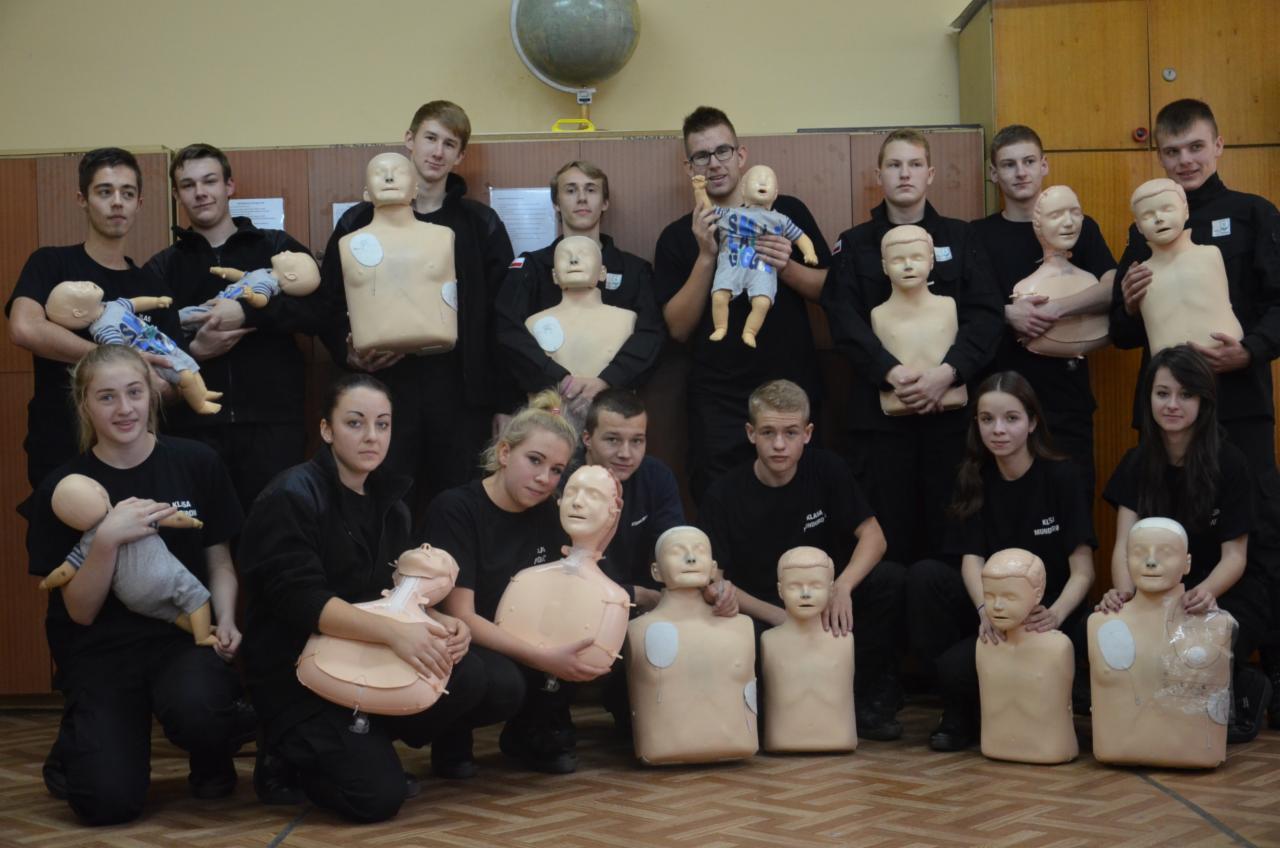 Grupa młodych ludzi w czarnych koszulkach pozuje z manekinami do ćwiczeń resuscytacji, w tym z manekinem niemowlęcia w ubranku.