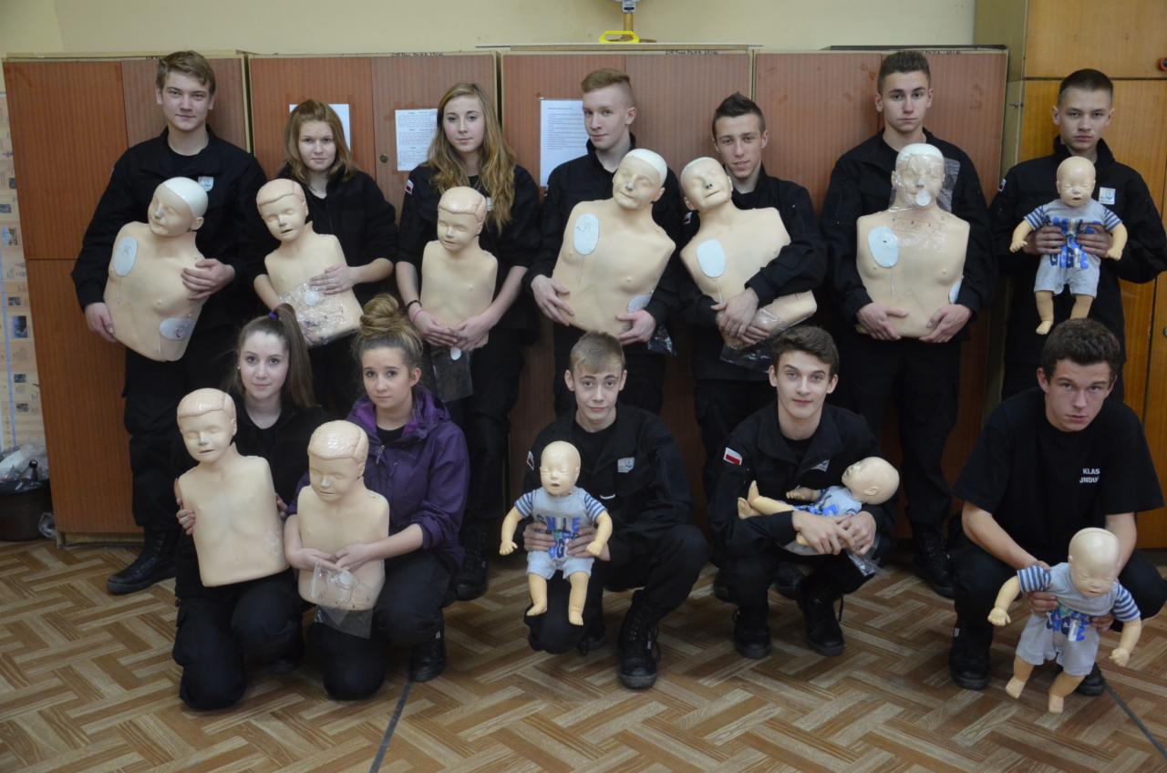 Grupa nastolatków w czarnych strojach pozuje z manekinami do nauki pierwszej pomocy, część z nich trzyma manekiny niemowląt w ubrankach, w tle szafki.