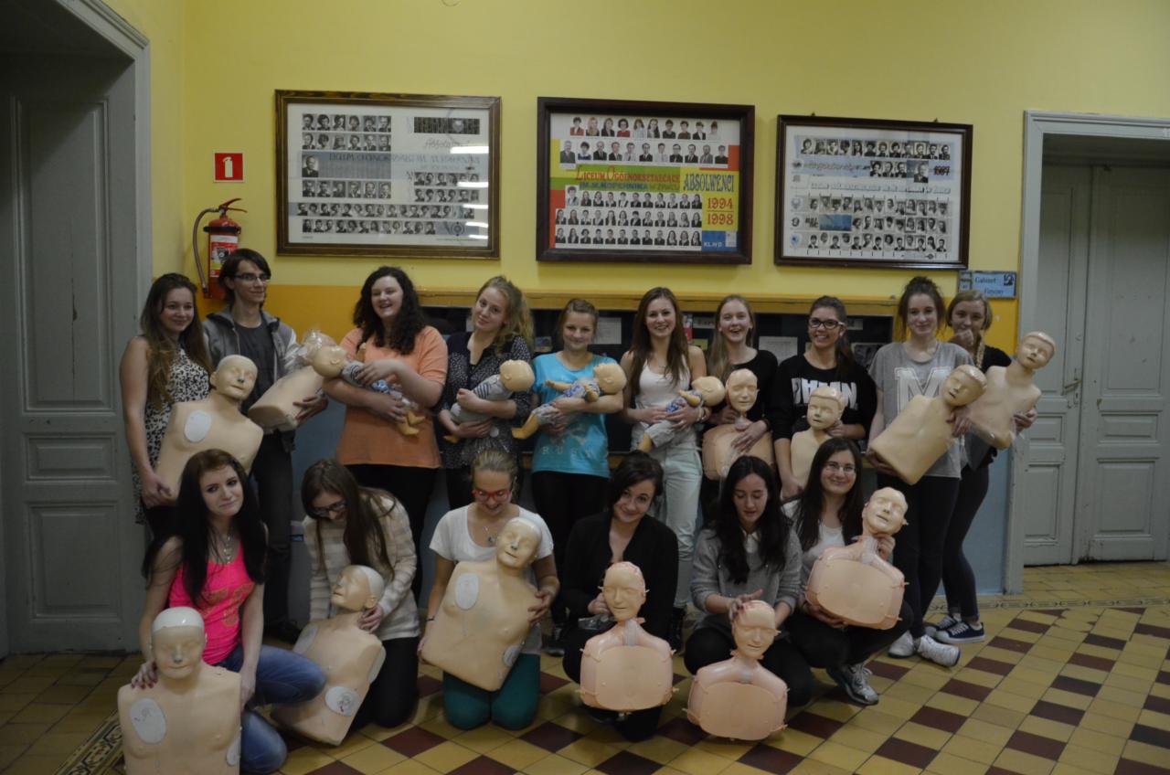 Grupa nastolatek pozuje z fantomami do resuscytacji w szkolnej sali, na ścianie wiszą oprawione zdjęcia klasowe.