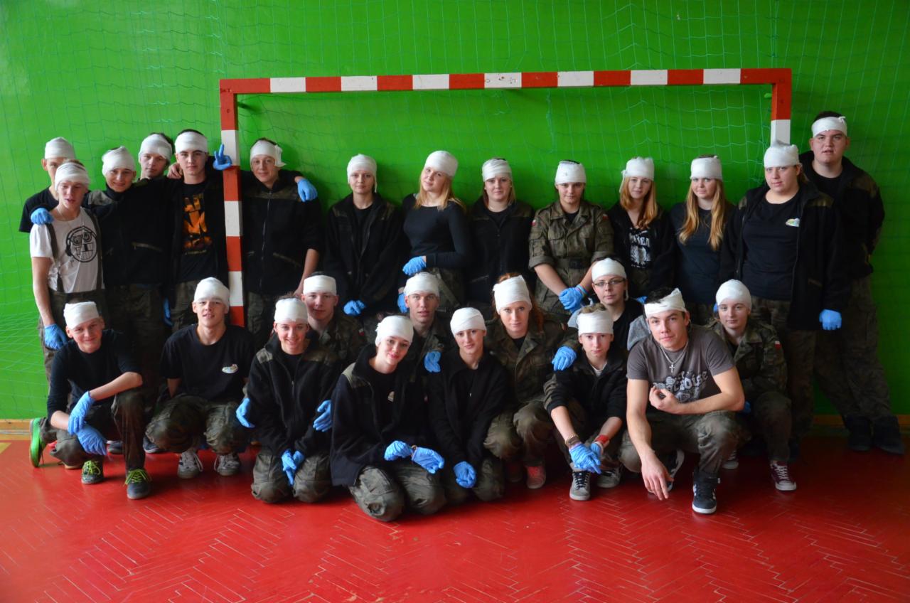 Grupa osób, część w mundurach, z opatrunkami na głowach i w niebieskich rękawiczkach, pozuje na tle zielonej siatki i bramki do piłki ręcznej po zakończonym szkoleniu.