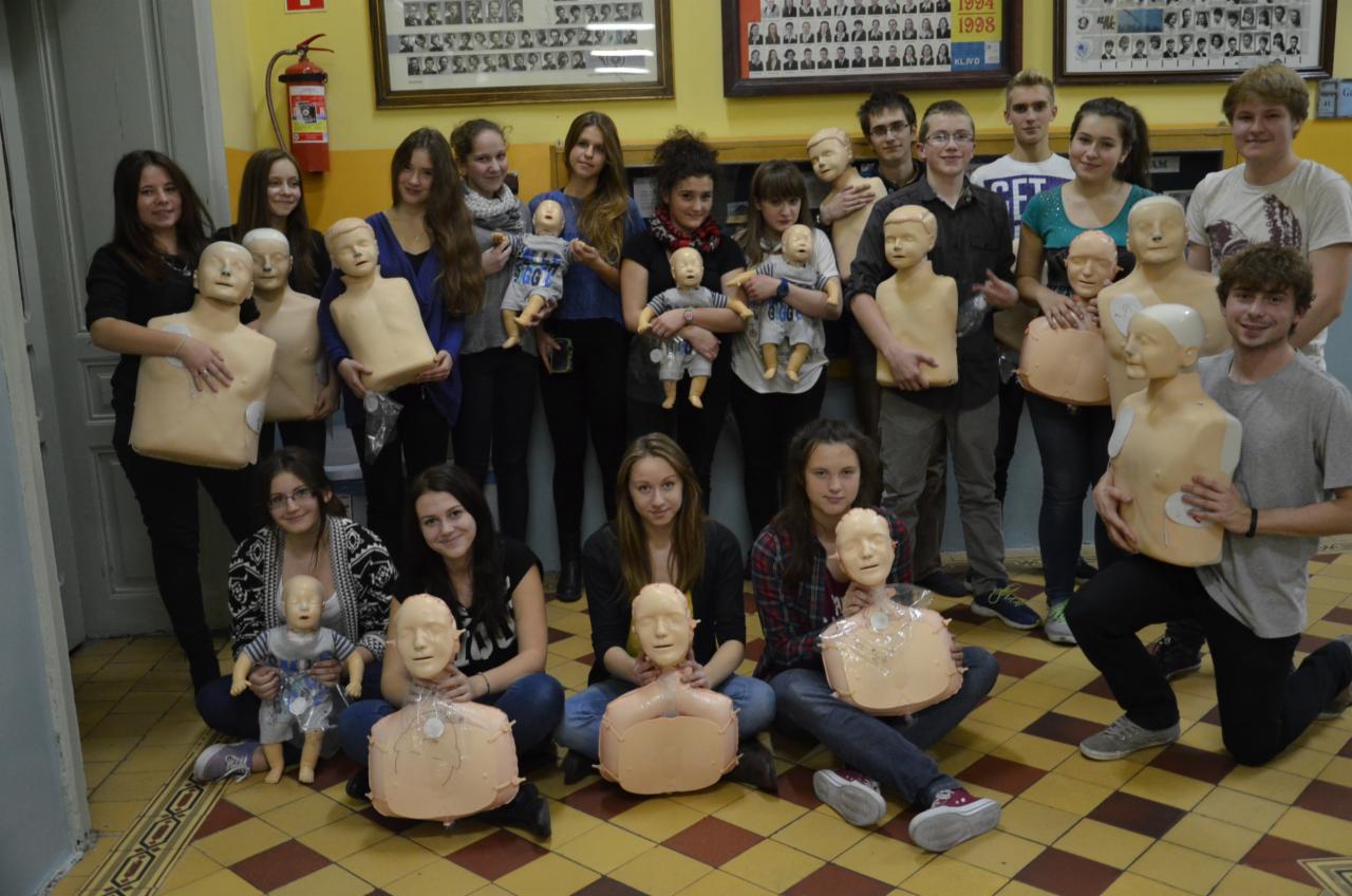 Grupa młodych ludzi, w tym kilka osób trzymających manekiny do nauki resuscytacji krążeniowo-oddechowej, pozuje do zdjęcia w sali z tablicami pamiątkowymi na ścianie i gaśnicą.
