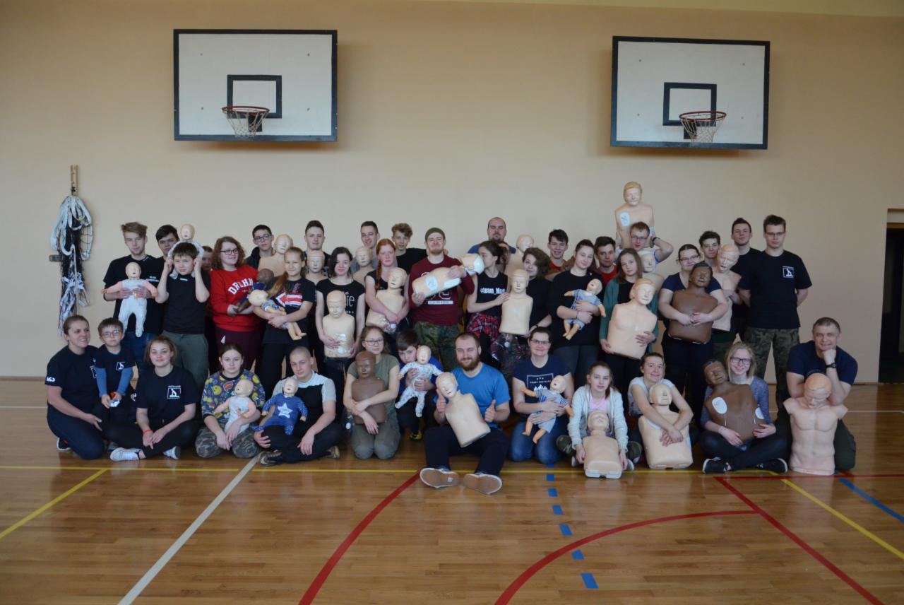 Grupa kursantów i instruktorów pozuje z manekinami do resuscytacji na sali gimnastycznej z koszami do koszykówki w tle.