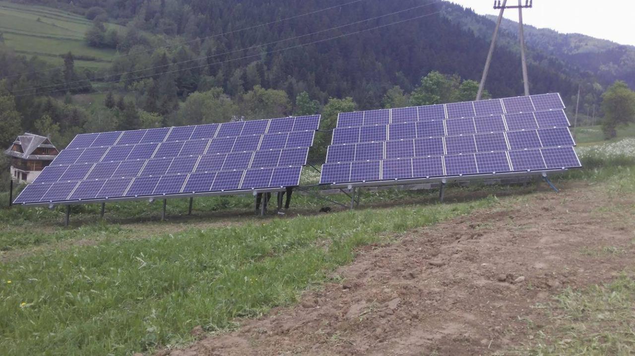 Dwa panele słoneczne na metalowej konstrukcji, zamontowane na trawiastym zboczu z widokiem na las i dom w tle.