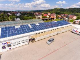 Duży budynek z logiem FedEx, na dachu instalacja z paneli słonecznych, szary samochód osobowy na parkingu, w tle zabudowa mieszkalna i zalesione wzgórze.