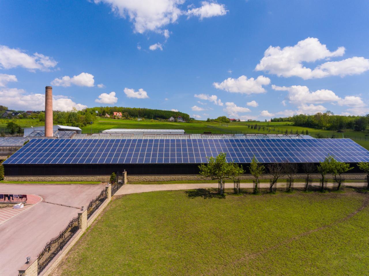 Rozległa instalacja fotowoltaiczna na dachu budynku gospodarczego z kominem w tle, widok z lotu ptaka.