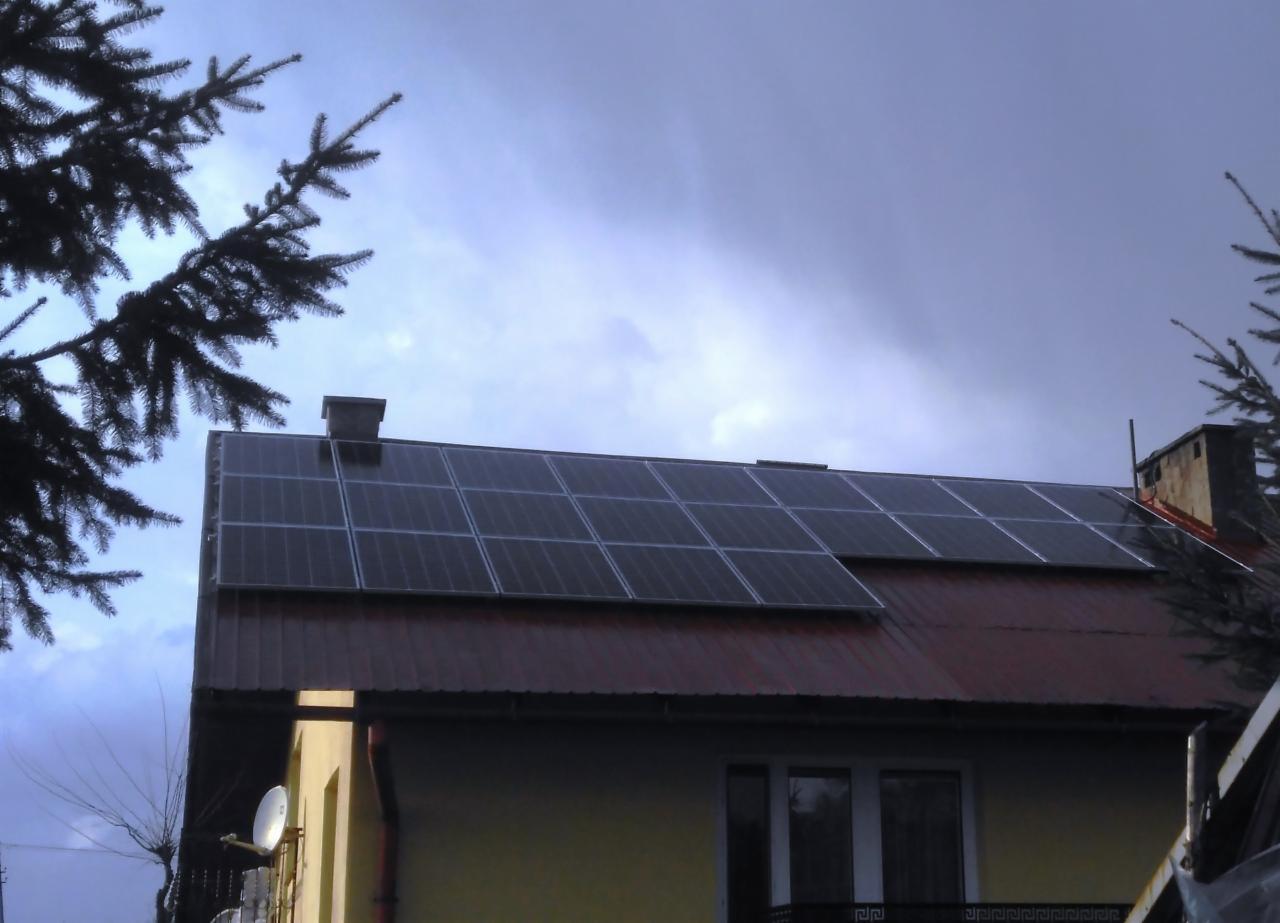 Panele słoneczne na czerwonym dachu domu jednorodzinnego, widok z dołu, pochmurne niebo w tle, gałęzie drzew po bokach kadru.