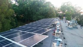 Rozległa instalacja paneli słonecznych na dachu z widocznymi elementami konstrukcyjnymi i transformatorami, otoczona zielenią drzew.