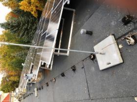 Panele słoneczne na dachu pokrytym papą, widoczne mocowania i instalacja elektryczna, drzewa w tle.