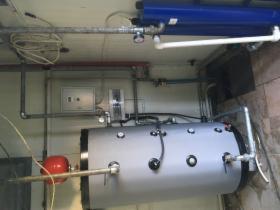 Szary, poziomy zbiornik buforowy z licznymi przyłączami hydraulicznymi i manometrami, zamontowany w pomieszczeniu technicznym z widoczną instalacją rurową pod sufitem i skrzynką elektryczną...