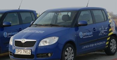 Dwa identyczne, niebieskie samochody osobowe marki Skoda Fabia z logo firmy na karoserii i napisem 'praca staje się bezpieczna' w żółto-czarne pasy, zaparkowane na zewnątrz w pochmurny dzień.