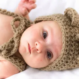 Noworodek leżący na białym tle, ubrany w dzierganą brązową czapeczkę z uszkami misia i dopasowany top na ramiączkach, z zaciekawionym spojrzeniem skierowanym w górę.