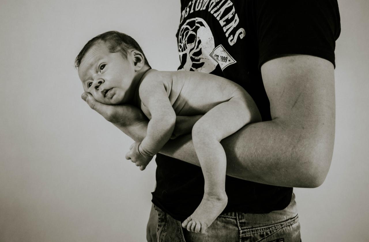 Noworodek w ramionach ojca, czarno-białe zdjęcie z delikatnym oświetleniem, dziecko spoczywa na przedramieniu, w tle widoczny fragment koszulki z nadrukiem i jeansy.