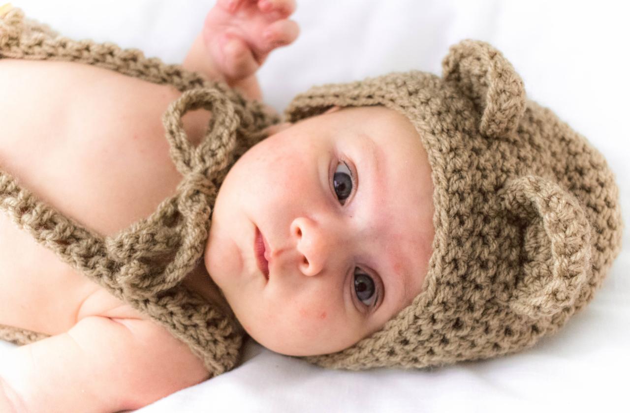 Noworodek leżący na białym tle, ubrany w dzierganą brązową czapeczkę z uszkami misia i dopasowany top na ramiączkach, z zaciekawionym spojrzeniem skierowanym w górę.
