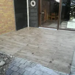 Wyłożony płytkami imitującymi drewno taras z widokiem na ogród przez przeszklone drzwi przesuwne.