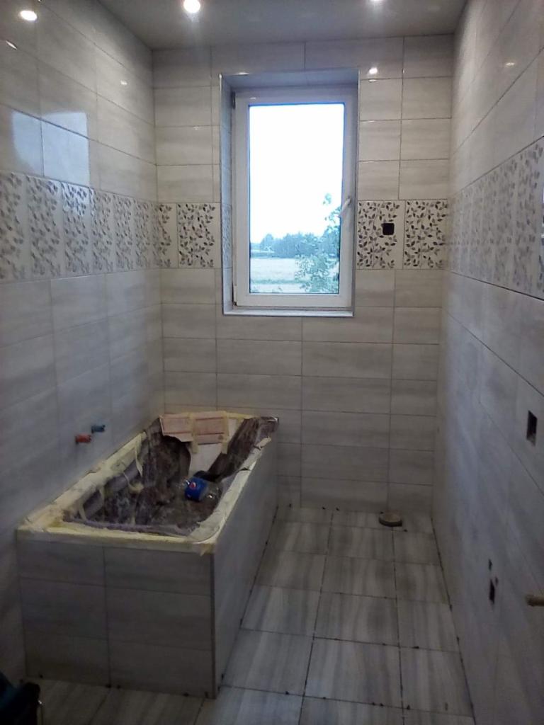 Łazienka w trakcie remontu: obudowana wanna przygotowana do wykończenia płytkami, ściany i podłoga częściowo wyłożone jasnymi, poziomymi płytkami z dekoracyjnym pasem, okno z widokiem na zieleń.