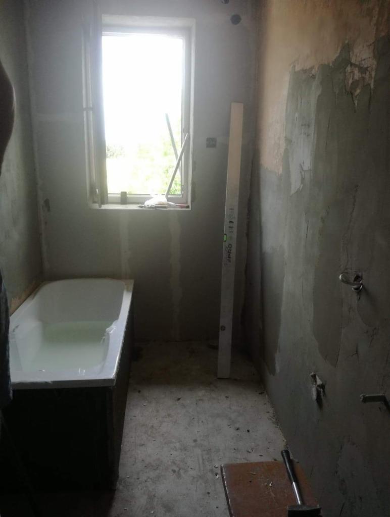 Łazienka w trakcie remontu w Wolazelichowskiej. Widoczna wanna wypełniona wodą, ściany w surowym tynku, poziomica oparta o ścianę i narzędzia na podłodze.