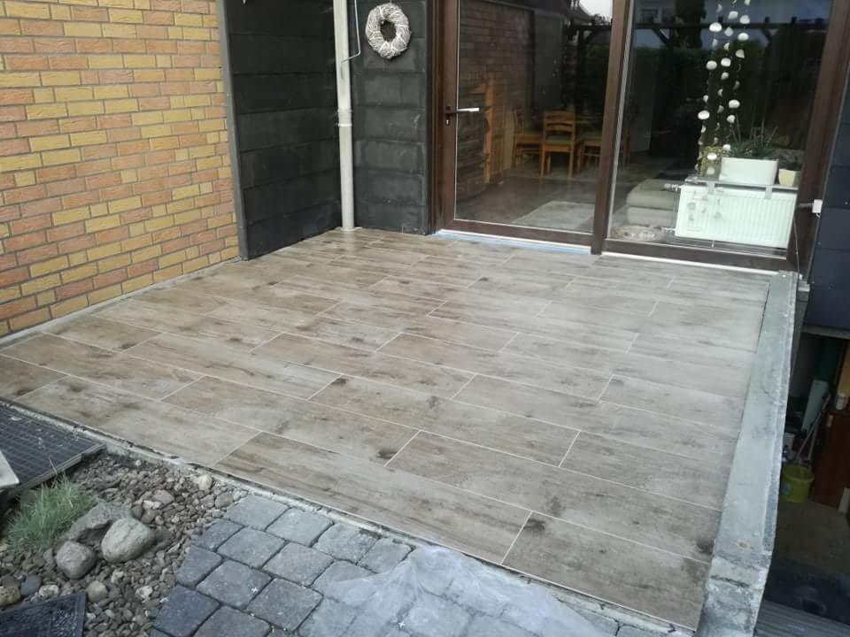 Wyłożony płytkami imitującymi drewno taras z widokiem na ogród przez przeszklone drzwi przesuwne.