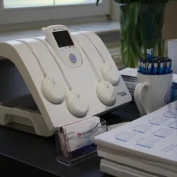 Białe urządzenie do elektrostymulacji mięśni z czterema elektrodami na czarnym stole, obok wizytówki, kalendarz i kubek z długopisami.