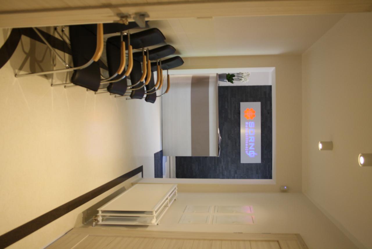 Poczekalnia z rzędem krzeseł z drewnianymi oparciami, widok na recepcję z logo firmy i dekoracyjnym kamieniem na ścianie.