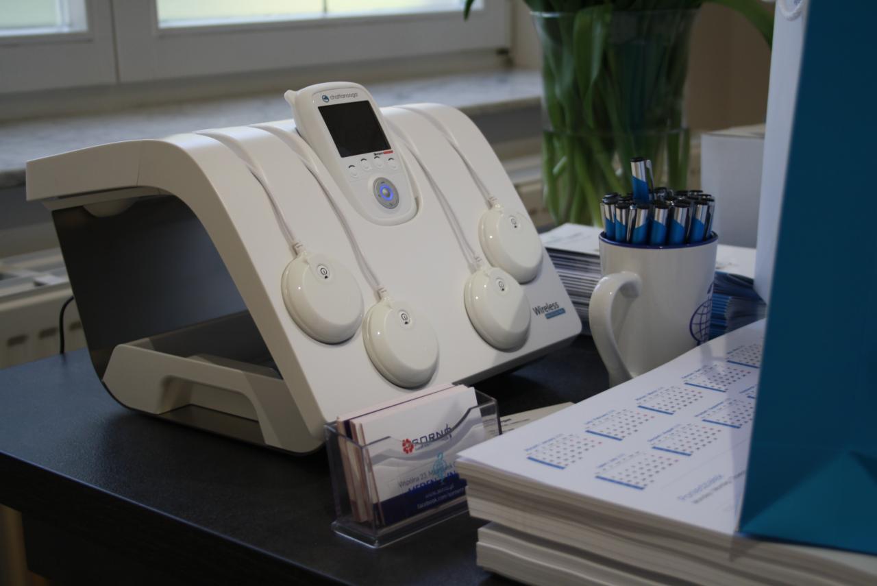 Białe urządzenie do elektrostymulacji mięśni z czterema elektrodami na czarnym stole, obok wizytówki, kalendarz i kubek z długopisami.