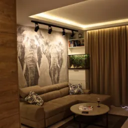 Nowoczesny salon z fototapetą przedstawiającą słonie, oświetleniem punktowym i narożną sofą w odcieniach brązu, uzupełniony akwarium i szklanką wina na okrągłym stoliku.