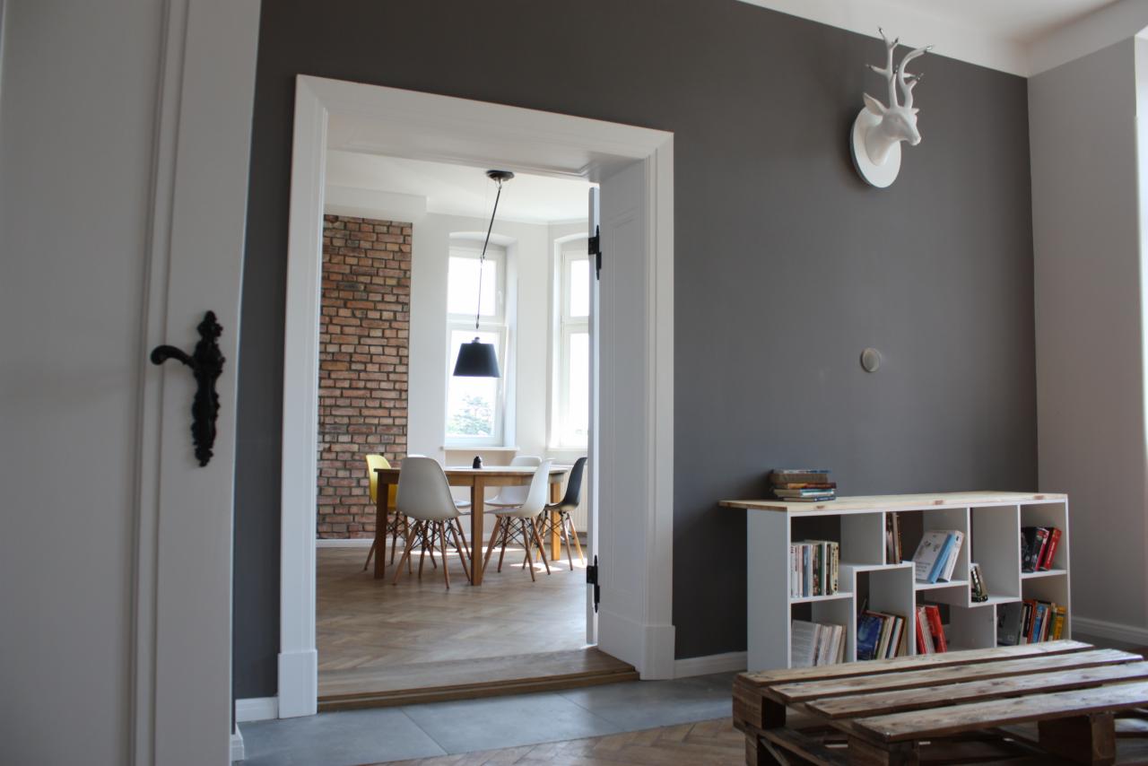 Nowoczesne wnętrze mieszkania z szarą ścianą, białym regałem z książkami i dekoracją głowy jelenia, widok na jadalnię z ceglaną ścianą przez otwarte drzwi z czarną klamką.