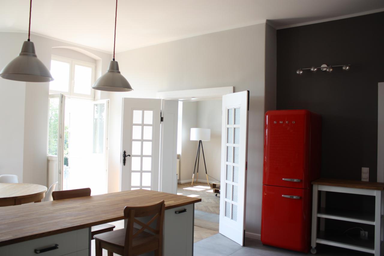 Jasna kuchnia z drewnianym blatem wyspy, otwartymi drzwiami balkonowymi i czerwona lodówką SMEG przy ciemnej ścianie.