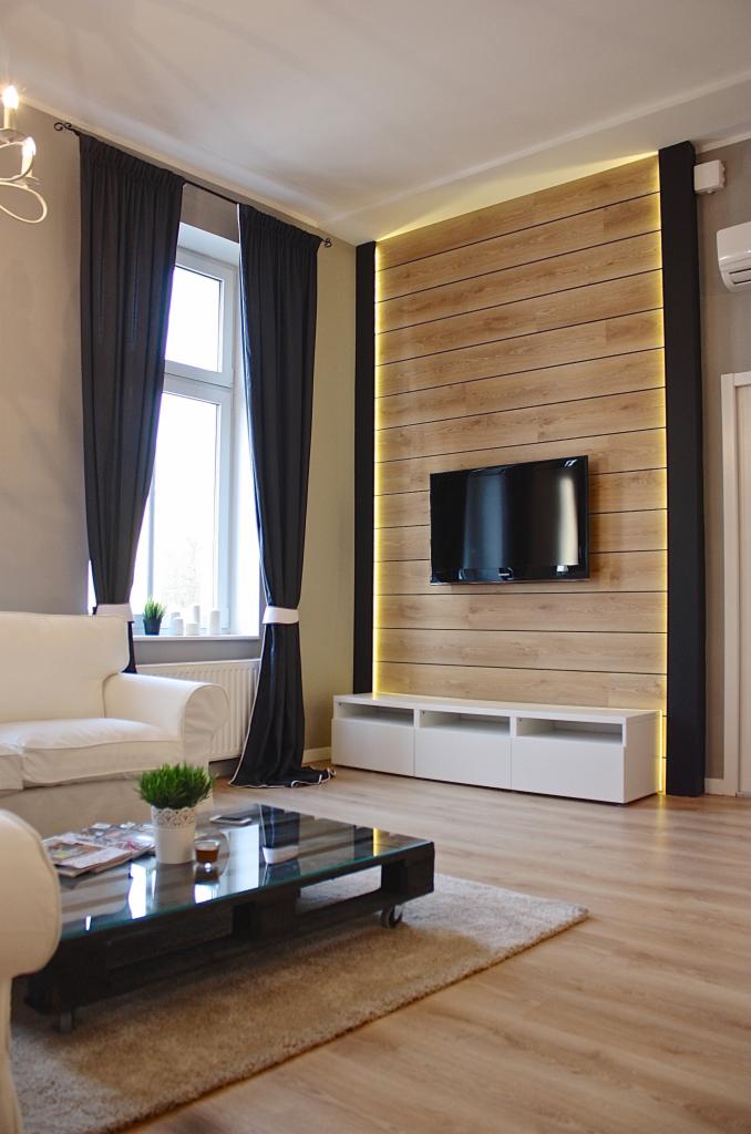 Nowoczesny salon z drewnianą ścianą podświetloną LED, telewizorem, białym regałem, ciemnymi zasłonami i stolikiem kawowym z palet.