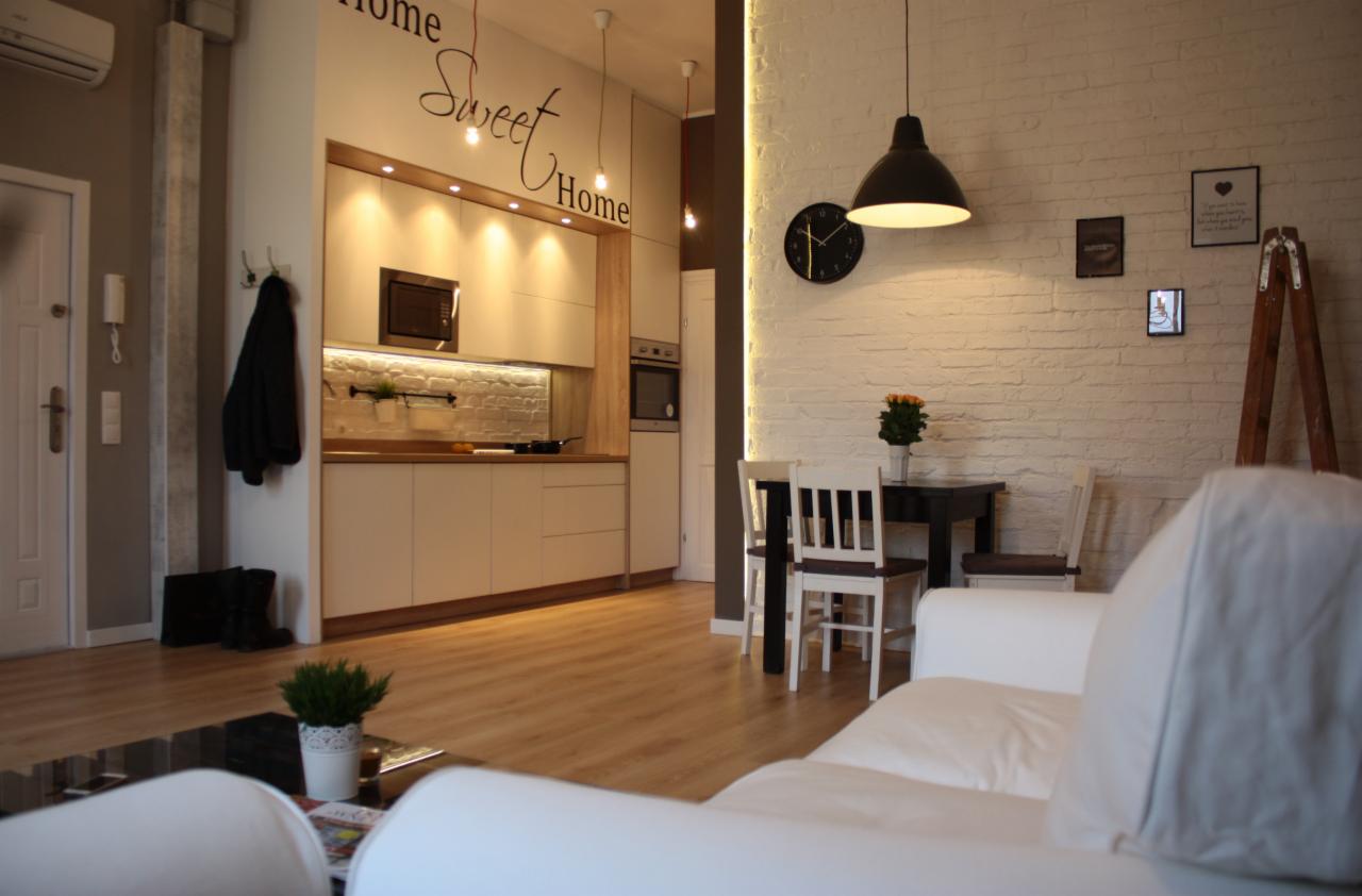 Jasne, przestronne wnętrze mieszkania z aneksem kuchennym, białymi meblami, ceglaną ścianą z dekoracjami i drewnianą podłogą, widoczne elementy dekoracyjne: zegar, kwiaty i napis 'Home Sweet Home'.