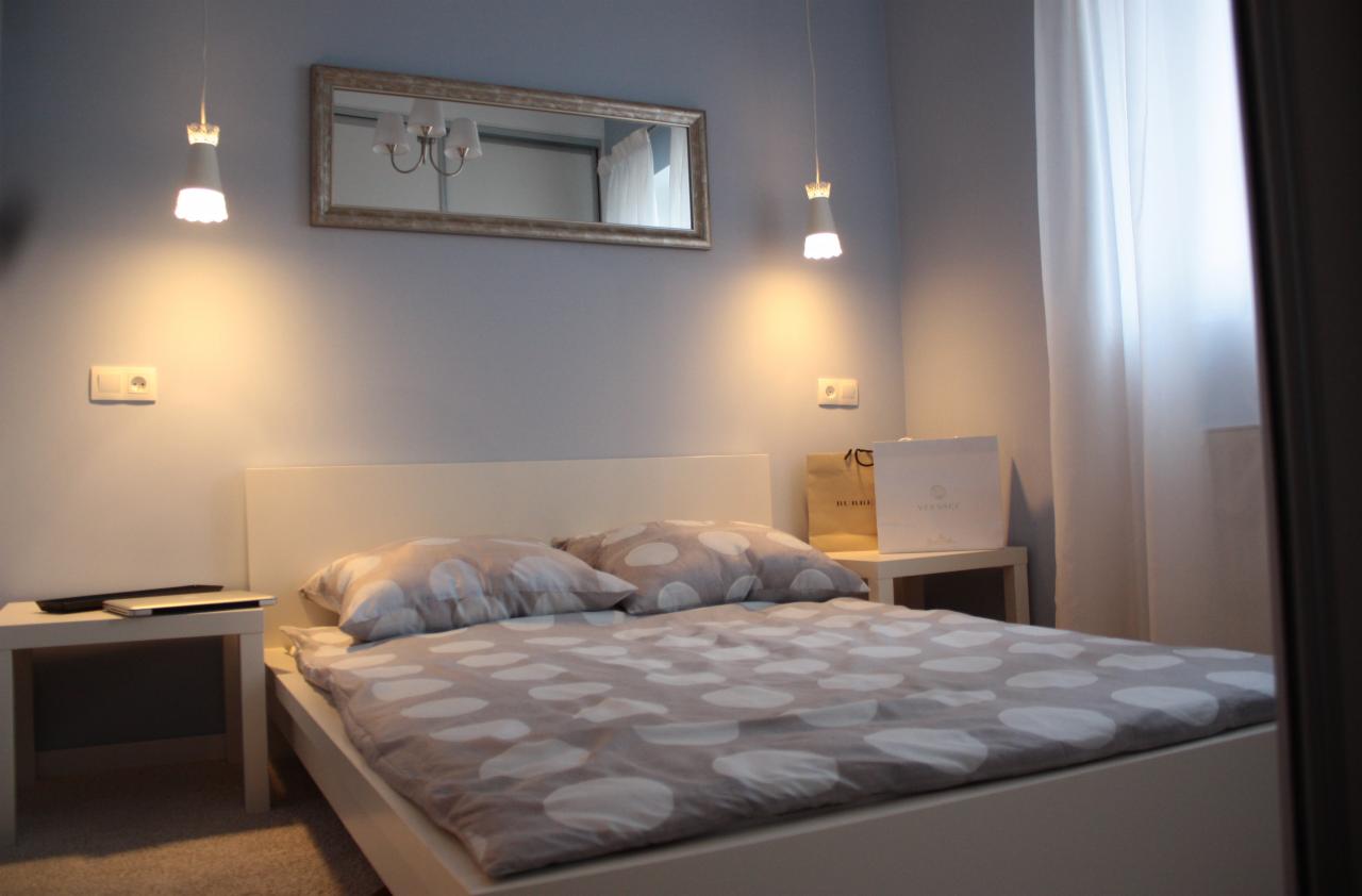 Nowoczesna sypialnia z białymi meblami, łóżkiem z szarą pościelą w białe grochy, lampkami wiszącymi i lustrem z odbiciem żyrandola, uzupełniona o torebki zakupowe na szafce nocnej.