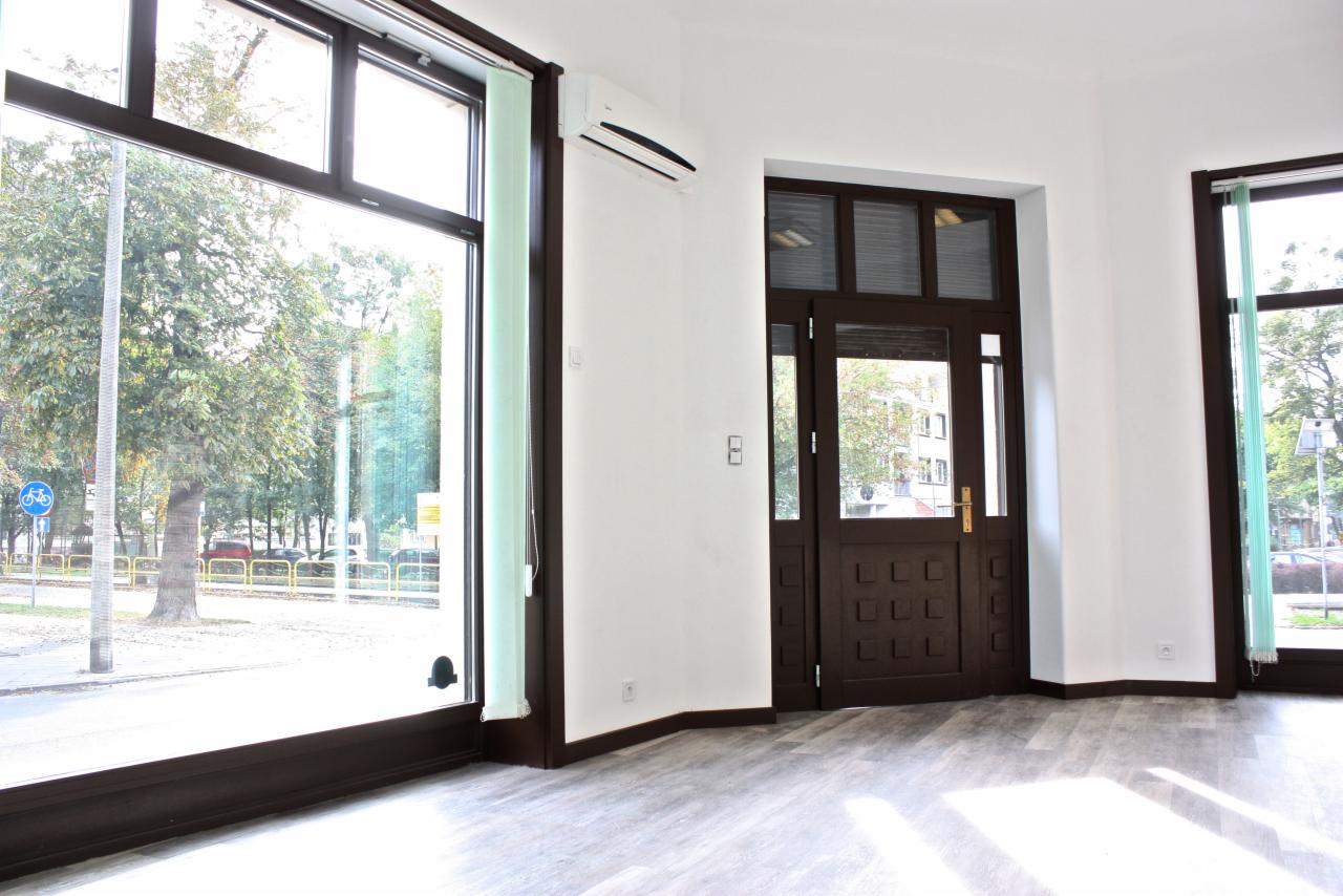 Wnętrze lokalu po remoncie: szare panele podłogowe, białe ściany, duże okna z widokiem na ulicę i drzewa, brązowe ramy okienne i drzwi, klimatyzator na ścianie.