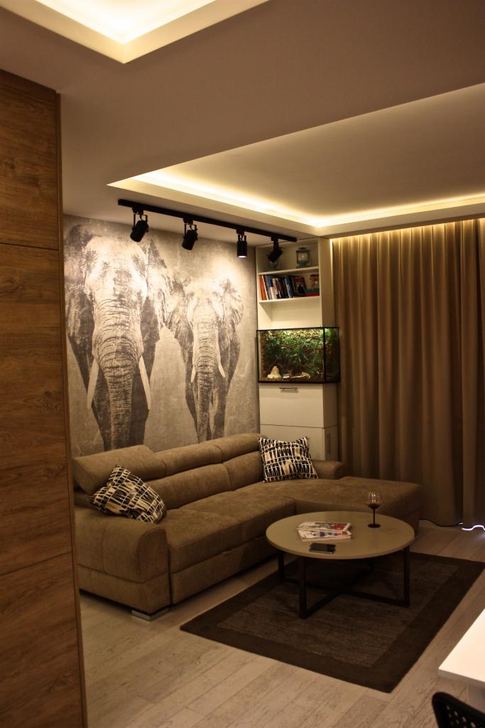 Nowoczesny salon z fototapetą przedstawiającą słonie, oświetleniem punktowym i narożną sofą w odcieniach brązu, uzupełniony akwarium i szklanką wina na okrągłym stoliku.