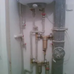 Szare rury kanalizacyjne i instalacja wodna z zaworami w pomieszczeniu w trakcie remontu, widoczne ślady po skuciu tynku na ścianie.