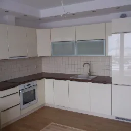 Jasna kuchnia z beżowymi, lakierowanymi frontami, ciemnym blatem i białymi płytkami na ścianie, widok na zabudowę kuchenną z piekarnikiem i zlewozmywakiem.