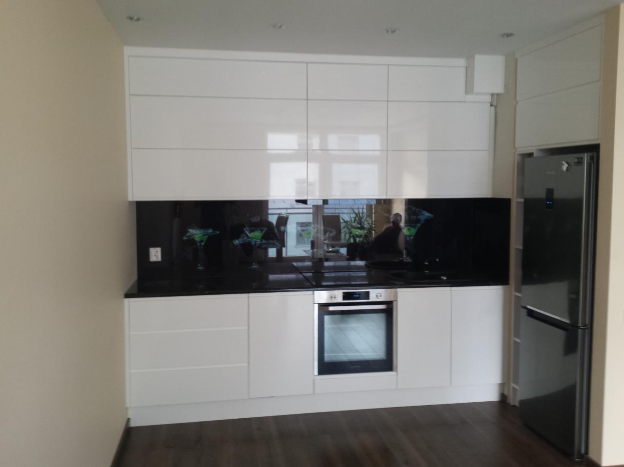 Biała zabudowa kuchenna z błyszczącymi frontami, czarnym blatem i panelem szklanym z motywem koktajli, widok na fragment podłogi z paneli.