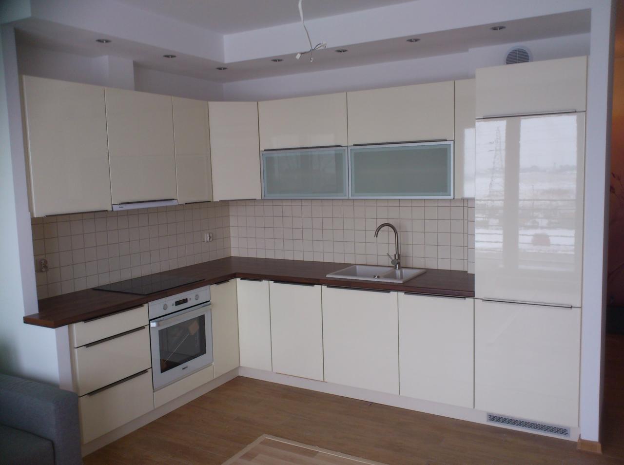 Jasna kuchnia z beżowymi, lakierowanymi frontami, ciemnym blatem i białymi płytkami na ścianie, widok na zabudowę kuchenną z piekarnikiem i zlewozmywakiem.