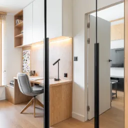 Nowoczesne biuro domowe z drewnianym biurkiem, szarą tapicerowaną krzesłem i białymi szafkami wiszącymi, oświetlone listwą LED, widoczne przez lustrzane drzwi przesuwne z czarnymi ramami.