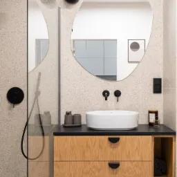 Nowoczesna łazienka z beżowymi płytkami lastryko, asymetrycznym lustrem, białą umywalką nablatową, drewnianą szafką pod umywalką z czarnymi uchwytami i czarną armaturą.