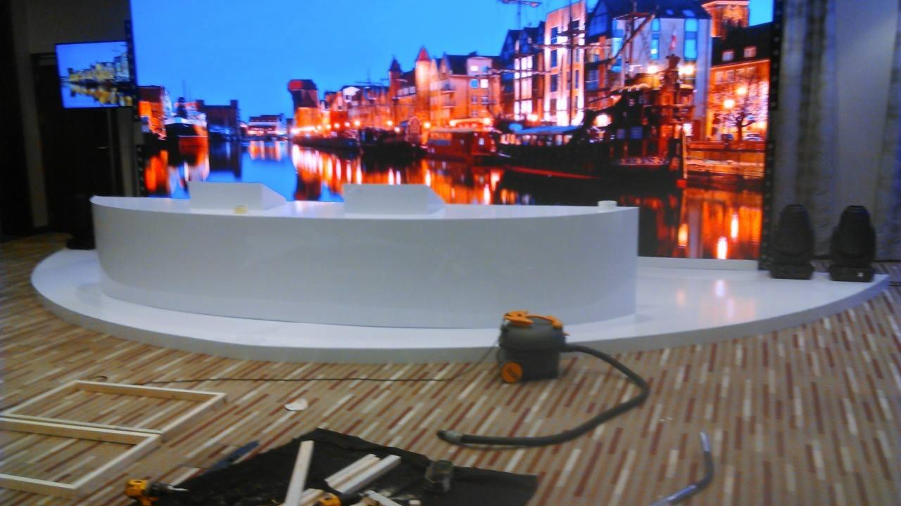 Biała, półokrągła konstrukcja sceniczna z tłem przedstawiającym nocny krajobraz miasta; na podłodze leżą drewniane ramy, narzędzia i odkurzacz.