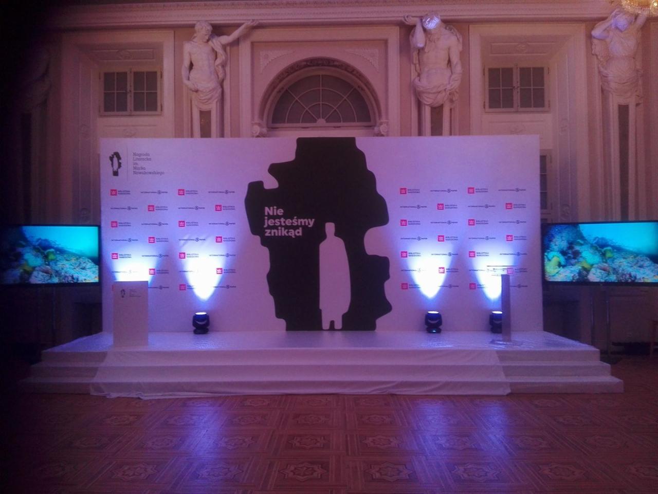 Scena z białym tłem z czarnym konturem postaci i napisem 'Nie jesteśmy znikąd', dwa monitory po bokach wyświetlające podwodny krajobraz, oświetlenie punktowe, dekoracyjne rzeźby na ścianie w tle.