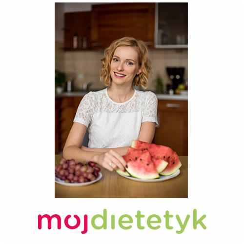 Uśmiechnięta blondynka w białej bluzce siedzi przy stole z talerzem arbuza i winogronami na tle kuchennych szafek.