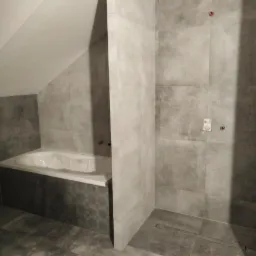 Szara łazienka w trakcie wykańczania, widoczna wanna pod skosem dachu, płytki imitujące beton na ścianach i podłodze, brak armatury, surowy stan.